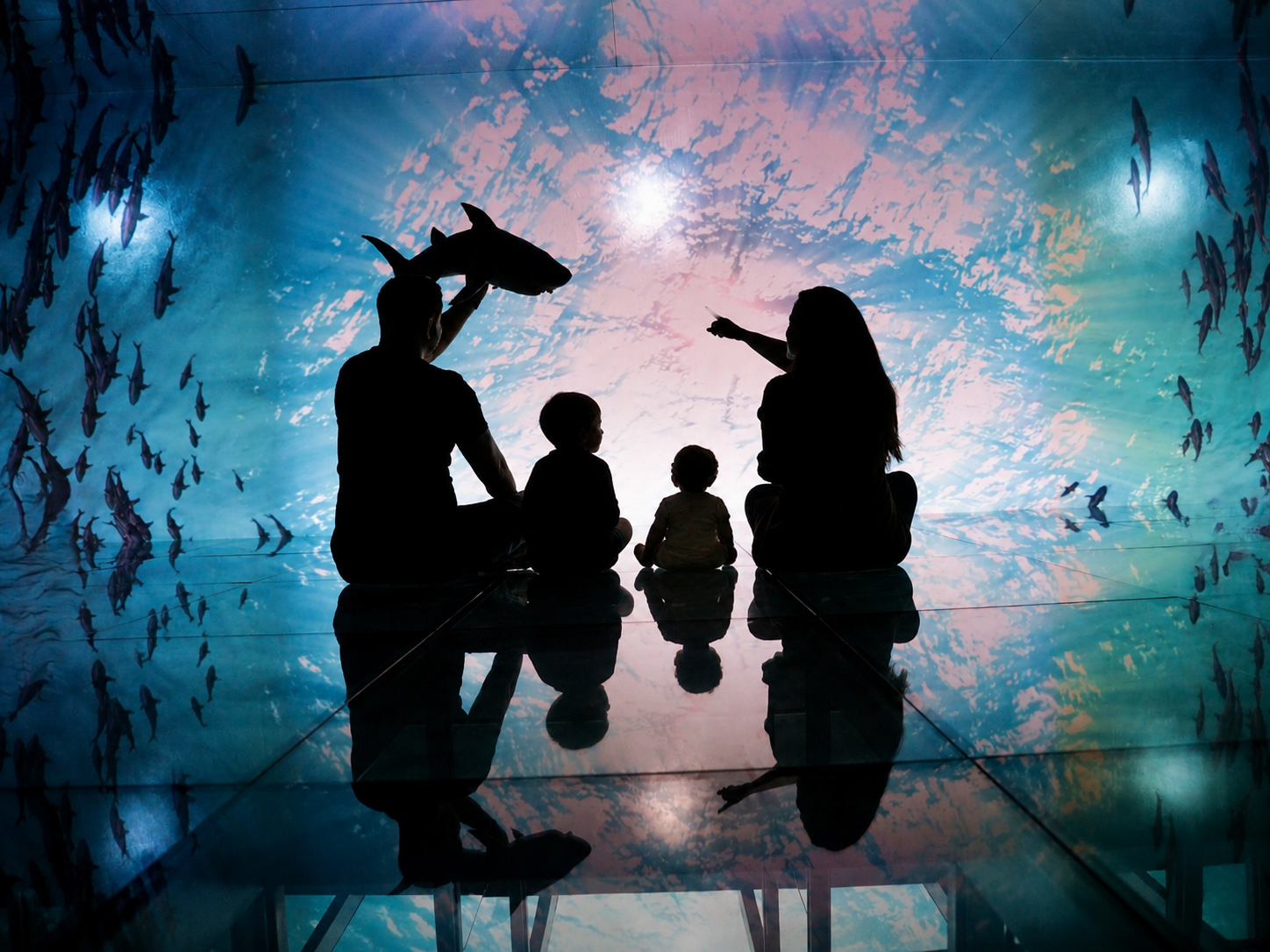 Silhouettes of a family sitting together inside an aquarium tunnel, watching light and fish move overhead